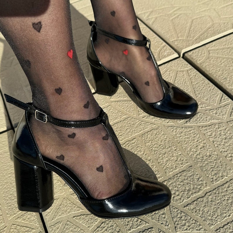May include: A pair of black heeled shoes with a t-strap design. The shoes are made of patent leather and have a chunky heel. The person wearing the shoes is wearing black sheer tights with a small red heart pattern.