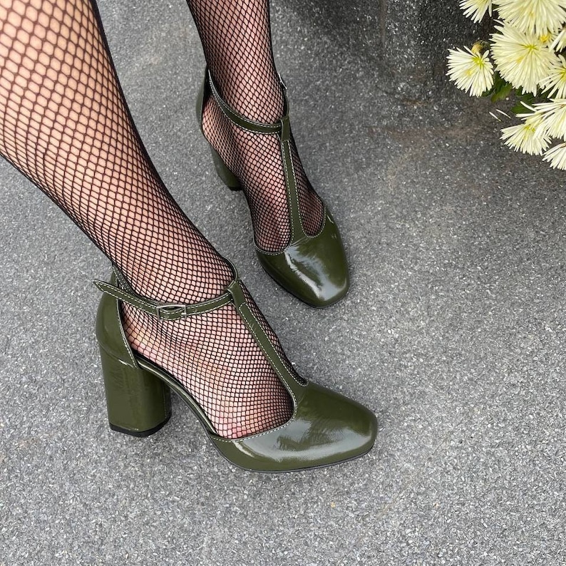 May include: A pair of green patent leather heeled shoes with a t-strap design. The shoes have a chunky heel and are worn with black fishnet tights.