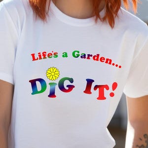 以下が含まれることがあります： 「Life's a Garden... DIG IT!」のカラフルな文字と黄色の花のグラフィックがプリントされた白いTシャツ。カジュアルな普段着です。