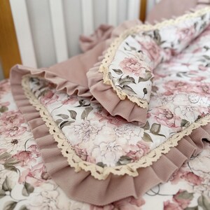 Könnte beinhalten: Ein Baby-Bettwäsche-Set mit einem rosa-weißen Blumenmuster. Das Set umfasst eine Decke, ein Nestchen und ein Kissen, alle mit einem Rüschenrand und Spitzenbesatz. Die sanften Farben schaffen eine zarte und gemütliche Atmosphäre.