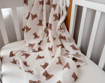 May include: A white muslin swaddle blanket featuring a repeating pattern of brown teddy bears. The fabric has a crinkled, textured appearance and is draped over the side of a white wooden crib, showcasing the soft, lightweight material.