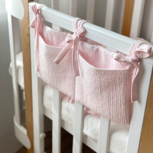 Può includere: Un organizer in tessuto rosa con due tasche è attaccato a una culla bianca. L'organizer è fissato con lacci rosa. La culla ha una struttura bianca con dettagli in legno.