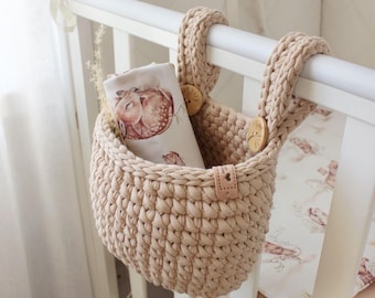 May include: A beige crochet basket with wooden buttons and leather tag hangs on a white crib. The basket holds a baby blanket with a watercolor illustration of a sleeping fawn. The nursery decor is soft and neutral.