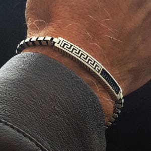 Könnte beinhalten: Silberfarbenes Armband mit griechischem Schlüsselmuster. Das rechteckige Mittelteil hat eine schwarze Einlage und das Band besteht aus Kettengliedern. Das Bild zeigt das Armband an einem Handgelenk vor dunklem Hintergrund.