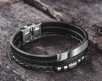 May include: A black leather bracelet with a stainless steel clasp and accents. The bracelet has a layered design with a braided leather band and a smooth, rectangular metal plate. The bracelet is displayed on a textured, dark surface.