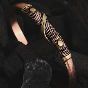 May include: Copper cuff bracelet with a woven mesh center and gold-colored accents. The bracelet features a decorative gold-colored snake design and two gold-colored rivets. The bracelet is set against a dark, textured background.