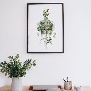 May include: A framed art print featuring a watercolor illustration of a human form filled with green foliage and white flowers. The artwork is set against a white background and framed in a black frame. The print is hanging on a white wall.