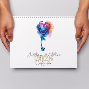 May include: A white spiral-bound calendar. The cover displays a watercolour illustration of a stethoscope shaped like a heart, with the text "Anatomy & Nature 2026 Calendar". The calendar is held by hands.