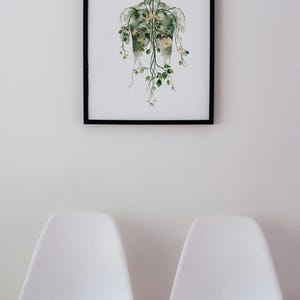 May include: A framed art print depicting a human form with botanical elements. The artwork features a black frame and a white background. The human form is illustrated with green veins and adorned with yellow flowers and green leaves. Two white chairs are in the foreground.