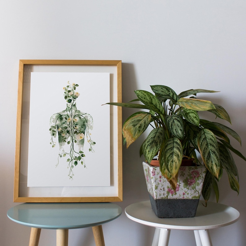 May include: A framed botanical print depicts a human form composed of plant life, with a wooden frame. A potted plant with green and yellow leaves sits nearby. The print and plant are on small, round tables.