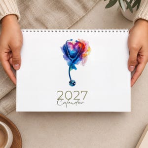 Può includere: Un calendario 2027 con rilegatura a spirale bianca che presenta un'illustrazione centrale ad acquerello di uno stetoscopio blu che forma un cuore con vivaci schizzi di vernice rosa, viola, gialla e blu. Il testo 2027 Calendar è stampato in grigio.