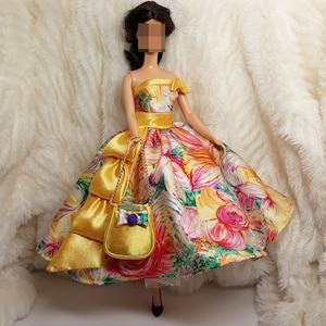 Könnte beinhalten: Eine Puppe trägt ein gelbes Kleid mit Blumenmuster und einem passenden gelben Satinband und einer Handtasche. Die Puppe hält die Handtasche mit einem goldenen Kettenriemen.