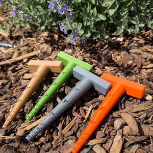 May include: Four colorful T-shaped plant markers in shades of tan, green, gray, and orange, each with inch measurements. The markers are arranged on a bed of wood chips, with a backdrop of green foliage and purple flowers.