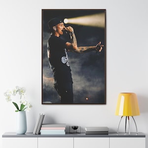 以下が含まれることがあります： マイクに向かって歌う、ステージ上のパフォーマーが写った額入りのキャンバスプリント。黒いTシャツ、黒いキャップ、黒いパンツを着用しています。背景は暗く、ステージ照明と煙があります。