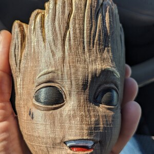 May include: A brown and gold 3D printed figurine of a character from the Guardians of the Galaxy film. The character has a large head with a wide smile and two black eyes. The figurine is made to look like wood.