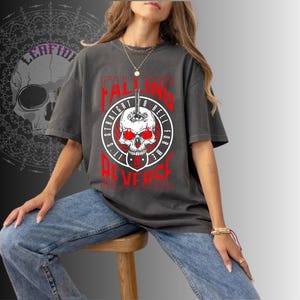 May include: Dark gray t-shirt with a graphic of a skull with red eyes and the text "I'M FALLING STRAIGHT TO HELL FOR ME IT'S NEVER". The shirt is worn with blue jeans.