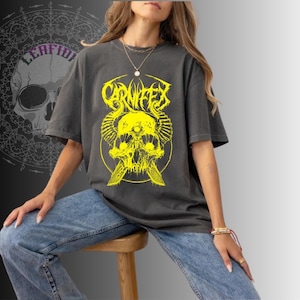 May include: Dark gray t-shirt featuring a yellow skull graphic with the word "CARNIFEX" above it. The design includes horns and crossed knives. The model is wearing blue jeans and sitting on a wooden stool.