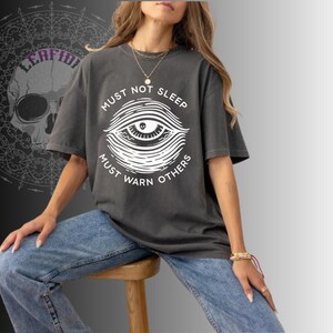 May include: Dark gray t-shirt with the text "MUST NOT SLEEP MUST WARN OTHERS" surrounding a white eye graphic. The shirt is worn with blue jeans. The overall theme is occult or spiritual.