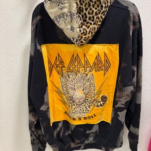 May include: A black hoodie with a bleached wash effect featuring a large yellow rectangular patch on the back. The patch displays a leopard illustration, the text Def Leppard, and Rock N' Roll. The hood has a split design with leopard print and paisley.