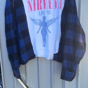 May include: A long-sleeve shirt featuring a white center panel with a Nirvana Live '93 graphic of a winged figure in blue and red text. The sleeves and side panels are made of a blue and black plaid fabric, creating a layered, reconstructed look.