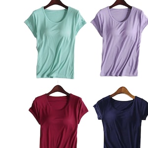 Op de afbeelding: Vier T-shirts met korte mouwen in verschillende kleuren: mintgroen, lavendel, bordeauxrood en marineblauw. Elk shirt heeft een ronde hals en hangt aan een houten hanger tegen een witte achtergrond. De shirts zijn gemaakt van zacht, licht materiaal.