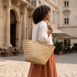 Könnte beinhalten: Eine große, naturfarbene geflochtene Strohtasche mit zwei geflochtenen Tragegriffen. Die Tasche hat ein breites, offenes Design mit einem strukturierten, horizontalen Webmuster über den gesamten Körper und bietet viel Platz für verschiedene Gegenstände.