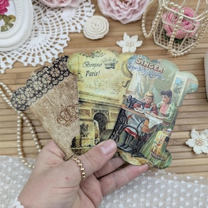 May include: Three vintage-style paper cards with scalloped edges. One card features a black lace design, another says "Bonjour Paris!" with an illustration of the Arc de Triomphe, and the third depicts a Singer sewing machine. The cards are held in a hand.