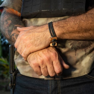 Puede incluir: Pulsera de cordón trenzado negro con detalles de madera y metal. La pulsera presenta dos cuentas rectangulares de madera y una cuenta metálica circular. La pulsera se lleva en la muñeca, con una camisa de camuflaje y vaqueros azul oscuro.