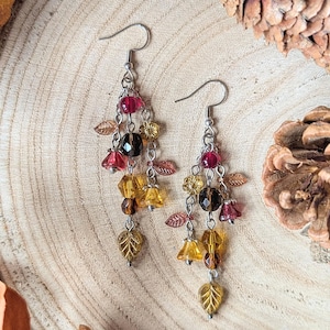 May include: A pair of drop earrings featuring silver-toned hooks and multiple strands of beads in shades of amber, brown, and deep red. The beads are shaped like small flowers and leaves, creating a layered, cascading design with metallic accents.