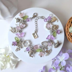 May include: A silver charm bracelet with various charms, including butterflies, flowers, hearts, and a small envelope. The bracelet features pink, purple, and green beads and charms. The bracelet is on a white surface.