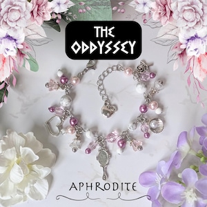 Può includere: Un braccialetto con ciondoli argentato con perline rosa e bianche, un ciondolo a forma di cuore e un piccolo specchio a mano. Il braccialetto è adornato da un ciondolo a conchiglia. La parola "APHRODITE" è sotto. Il testo "THE ODYSSEY" è in una scatola nera.