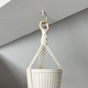 Peut inclure: Un pot en céramique blanc suspendu à un porte-plante en macramé avec un anneau en bois. Le porte-plante est accroché à un crochet au plafond.