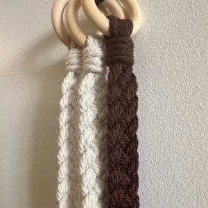 Op de afbeelding: Drie macramé plantenhangers met houten ringen. Twee zijn wit en één is bruin.