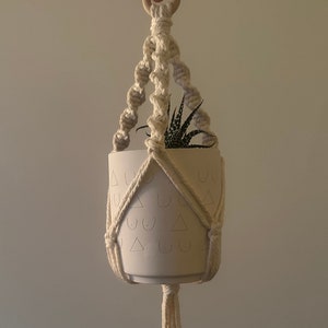 May include: A white ceramic planter with a pattern of triangles and breasts hangs from a macrame plant hanger. The hanger is made of natural cotton rope and has a wooden ring at the top.