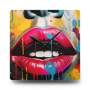 May include: A square wall clock with a colorful abstract design featuring a large pair of red lips with white teeth. The background is a mix of yellow, blue, and black paint splatters.