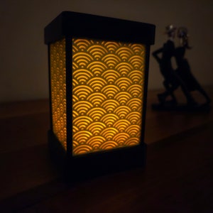May include: A black lamp with a geometric pattern on the front. The lamp is lit and the pattern is illuminated in a warm yellow color.