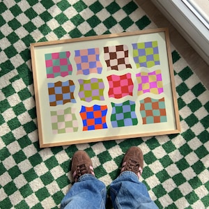 以下が含まれることがあります： ピンク、青、緑、茶色の色合いで、カラフルな波状のチェッカーボードパターンが並んだ額入りのアートプリント。アートワークは、緑と白のチェック柄のラグの上に置かれています。茶色のスニーカーと青いジーンズを履いた人の足が見えます。