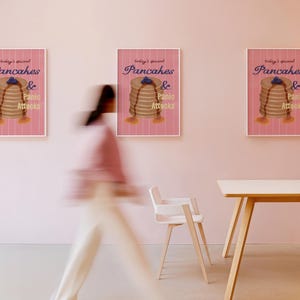 Puede incluir: Tres carteles enmarcados en una pared rosa. Cada cartel presenta una pila de panqueques con jarabe y arándanos, con el texto "today's special Pancakes & Panic Attacks". Una persona pasa por una silla y una mesa blancas.