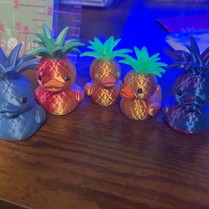 May include: Five 3D-printed pineapple duck figurines in various colors. The ducks have textured bodies and pineapple tops in blue, red, orange, and gold. The ducks are sitting on a wooden surface.