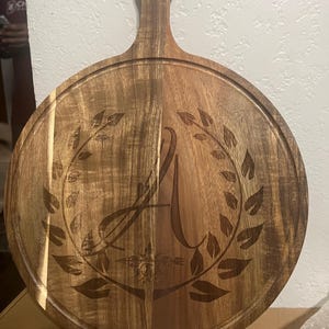 May include: A round wooden cutting board with a handle. The board features a carved design of the letter "A" within a laurel wreath. The wood has a natural grain and a dark brown color, with a groove around the edge.