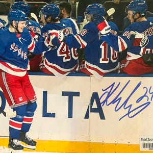 Può includere: Un giocatore di hockey con una maglia blu, rossa e bianca dei New York Rangers con il numero 24. Il giocatore sorride e tiene in mano una maglia. Altri giocatori sono visibili sullo sfondo. La parola "DELTA" è visibile.