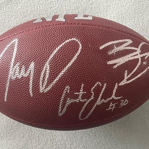May include: A maroon leather football with white signatures. The NFL logo is visible. The football has multiple white signatures on the surface. The football is a standard size and shape.