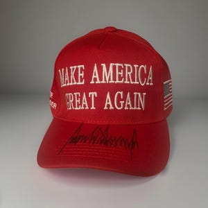 May include: A bright red baseball cap featuring the white embroidered text MAKE AMERICA GREAT AGAIN on the front crown. The brim displays a black signature. A small American flag is embroidered on the side, and the text TRUMP PENCE is on the side.