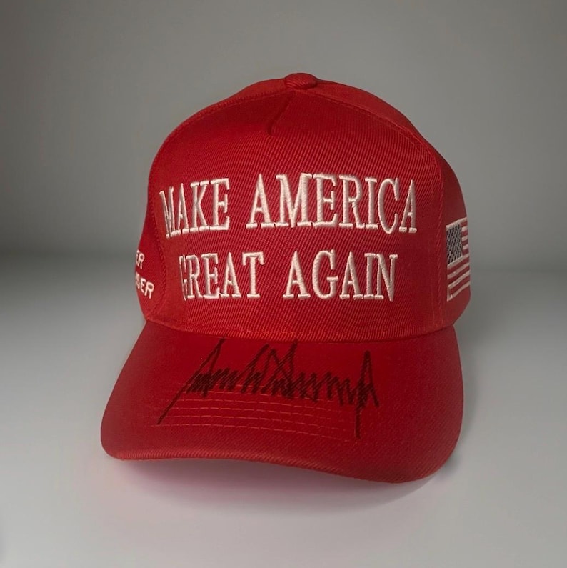 May include: A bright red baseball cap featuring the white embroidered text MAKE AMERICA GREAT AGAIN on the front crown. The brim displays a black signature. A small American flag is embroidered on the side, and the text TRUMP PENCE is on the side.