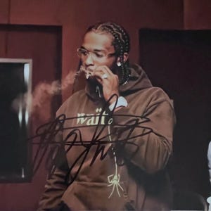 May include: An autographed brown hoodie with the word "wait" and a spider graphic. The person in the image is wearing glasses and smoking. The image is signed with a black marker.