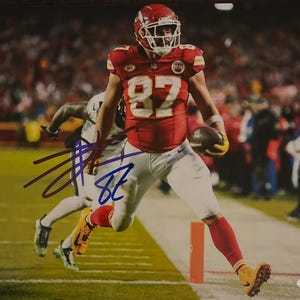 Puede incluir: Un jugador de fútbol americano con una camiseta roja y blanca con el número 87, corre con el balón. El jugador está firmando su nombre en tinta azul sobre un fondo blanco.