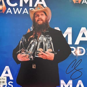 Könnte beinhalten: Eine Person hält drei Auszeichnungen aus klarem Glas mit goldenen Medaillons. Die Person trägt einen schwarzen Anzug und einen Cowboyhut. Der Hintergrund ist ein blaues Banner mit dem Text "CMA Awards". Eine Unterschrift ist rechts sichtbar.
