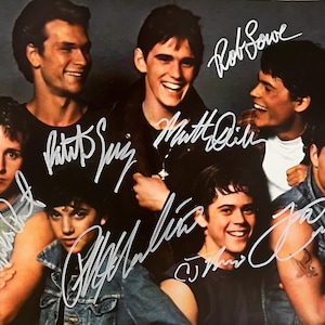 May include: A promotional photograph of the cast of The Outsiders featuring seven young actors with white ink signatures overlaid on the image. The group includes Patrick Swayze, Matt Dillon, Rob Lowe, Emilio Estevez, and Tom Cruise.