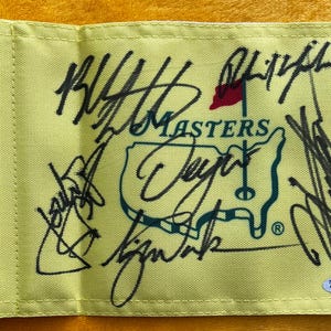 May include: A pale yellow Masters golf flag featuring a central green outline of the United States with a red flag icon. The fabric is covered in multiple black ink signatures and includes a small oval Total Sports Authentics sticker in the corner.