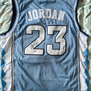 Może przedstawiać: Jasnoniebieski koszulka do koszyk&oacute;wki z napisem "JORDAN" i numerem "23" w kolorze białym. Koszulka ma podpis na numerze 23. Koszulka ma niebiesko-biały wz&oacute;r romb&oacute;w po bokach.
